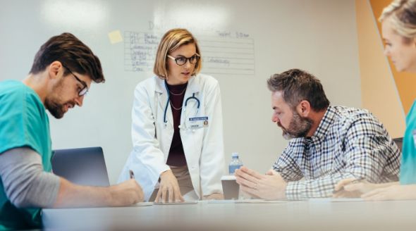 A doctor in a white coat stands at a meeting table with others seated